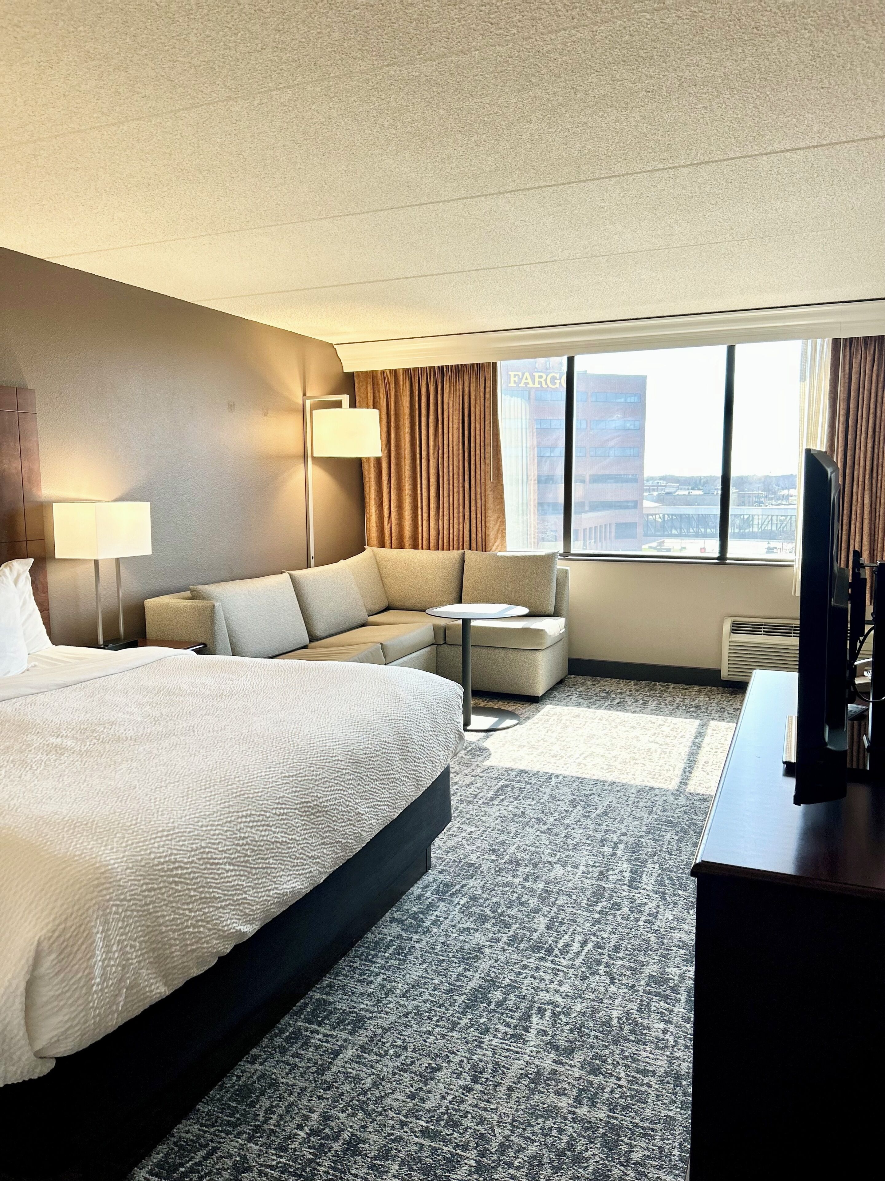 Ein Hotelzimmer mit einem großen Bett, einer Couch, einem kleinen Tisch, einem Fernseher und Blick auf die Stadt.