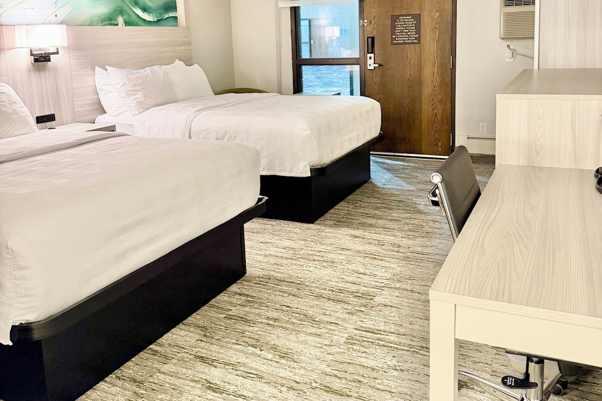 Une chambre d’hôtel avec deux lits, un bureau, une chaise, un tableau au mur et une porte avec une pancarte.