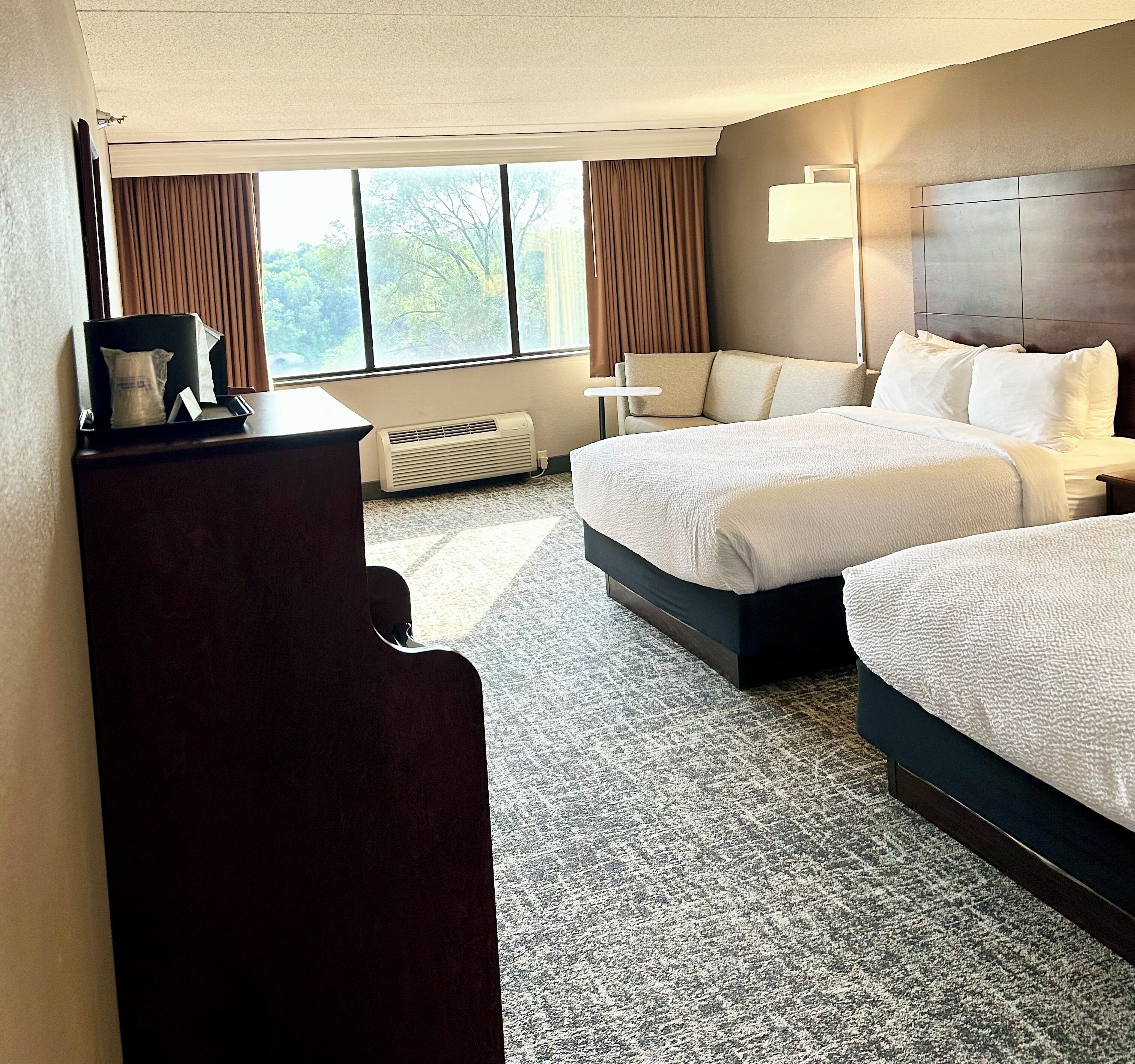 A hotel room with two beds, a desk, a chair, a lamp, a heater, and a window with curtains.