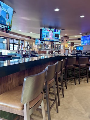 A bar area with multiple screens displaying sports content, bar stools, and a counter with a variety of bottles.