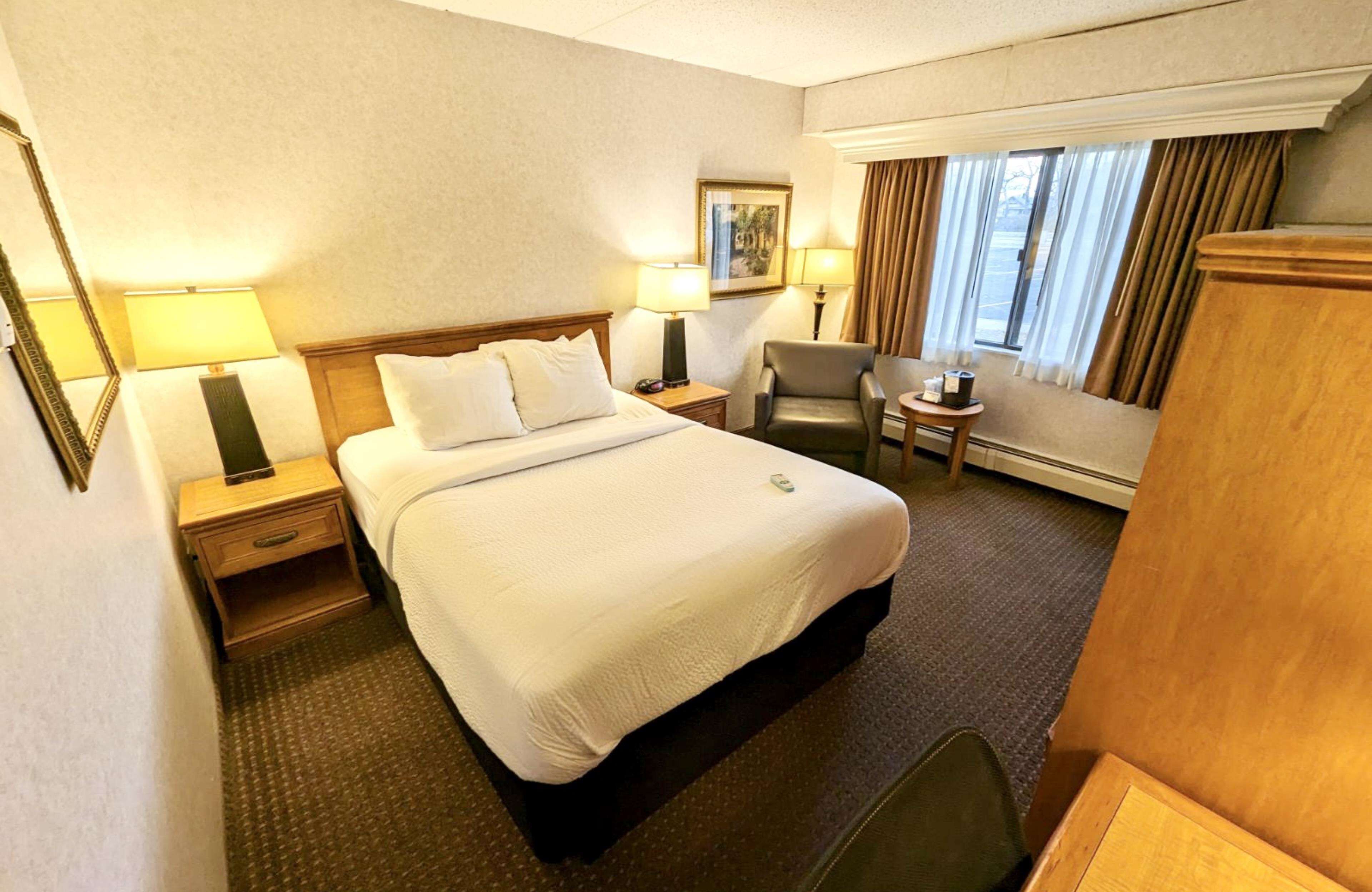 A hotel room with a large bed, two bedside tables, a chair, a small table, and a window with curtains.