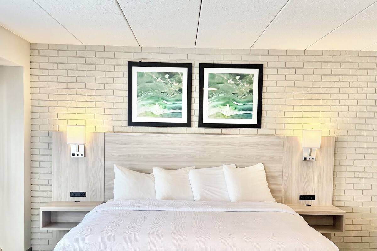 Habitación de hotel con una cama grande, dos mesitas de noche fijadas a la pared, un muro de ladrillo blanco y dos cuadros enmarcados sobre la cama.