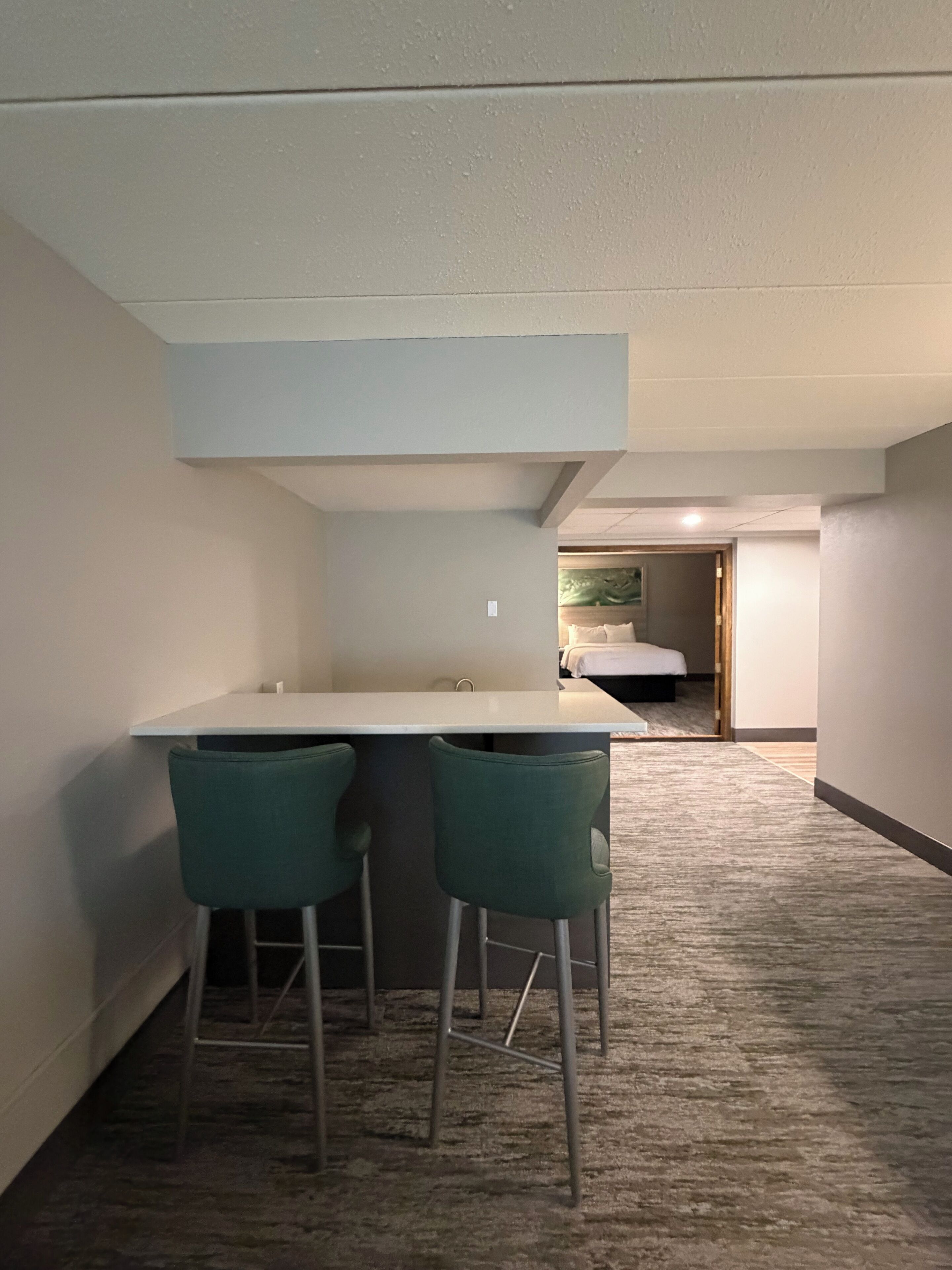 A hotel room with a kitchenette, two bar stools, a bed, and a carpeted floor.