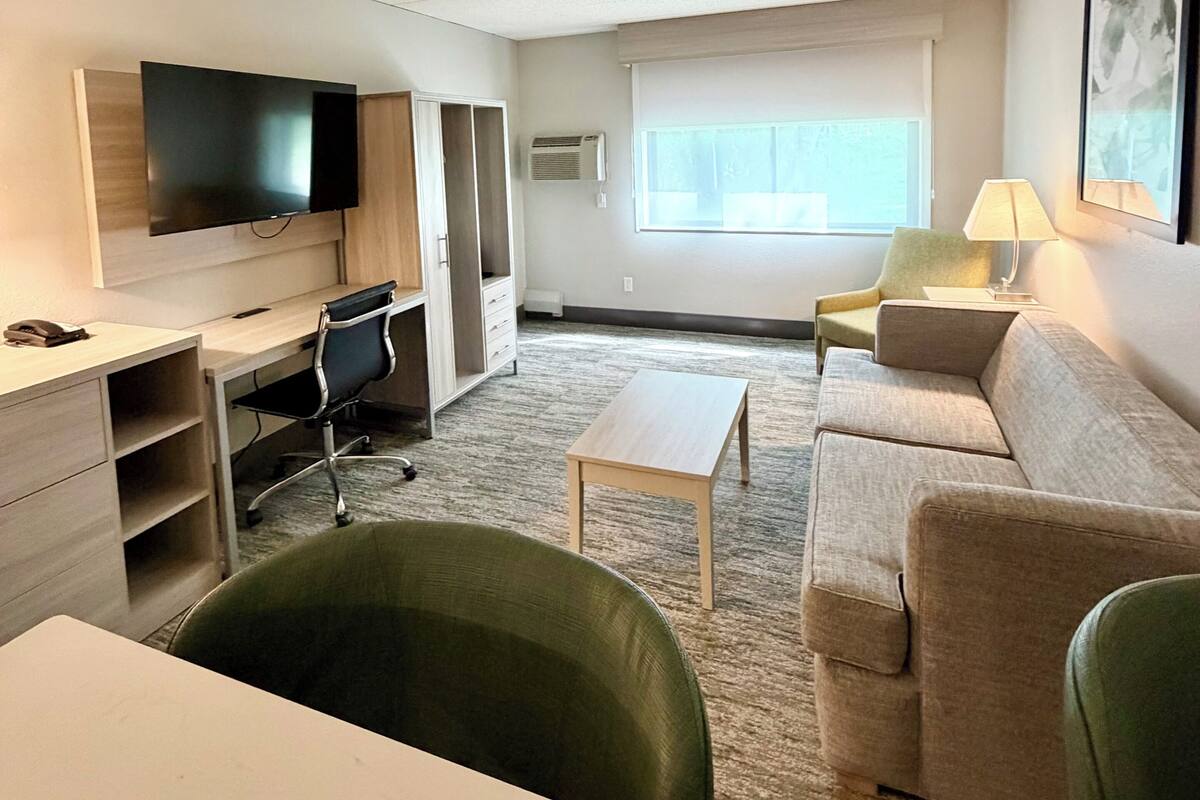Habitación de hotel con televisor de pantalla plana, escritorio, silla verde, sofá, mesita y ventana con vistas.