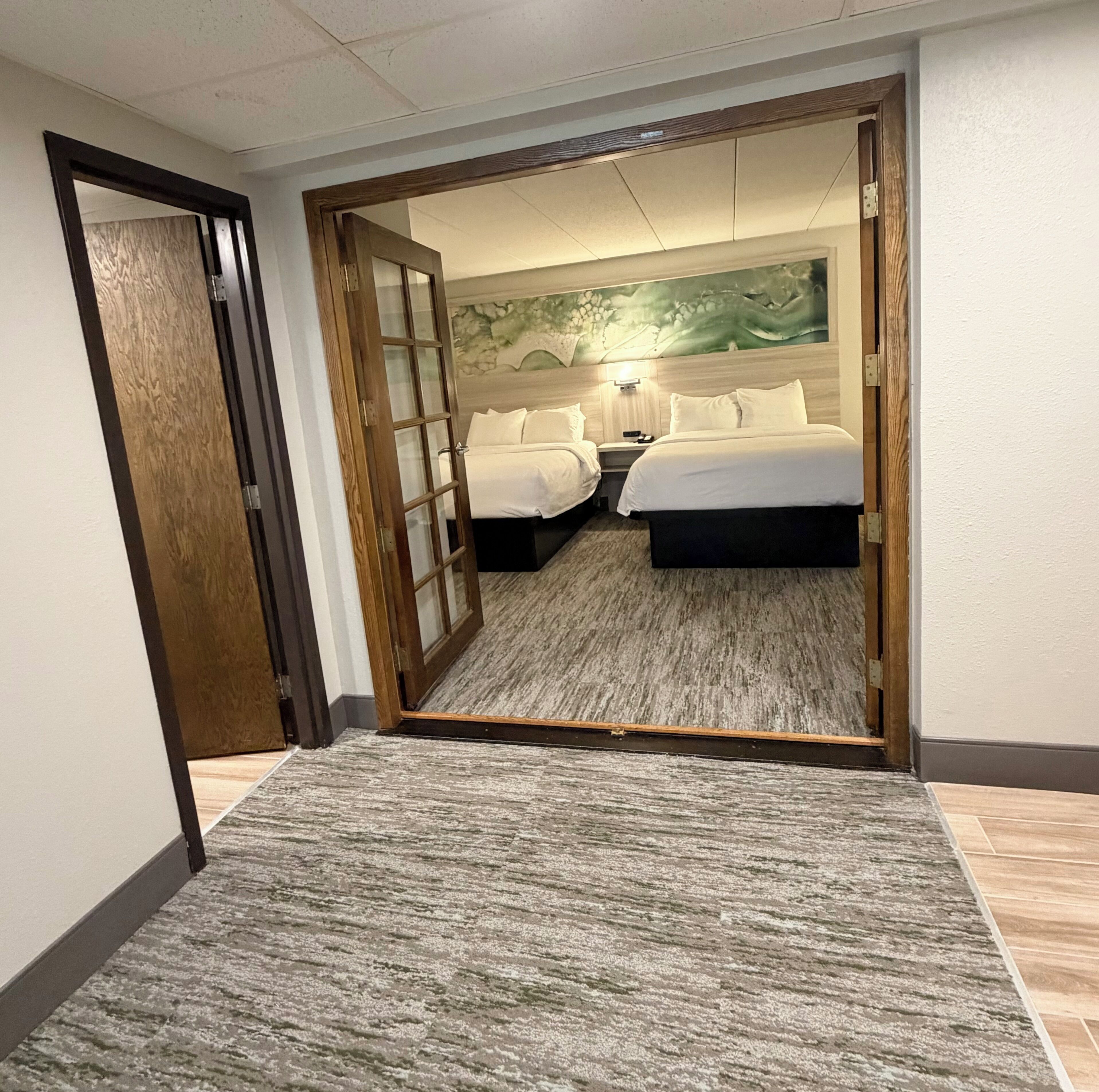 Une chambre d’hôtel avec deux lits, une porte en bois, un sol recouvert de moquette et un mur orné d’un motif abstrait vert.