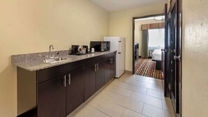 A compact kitchen with a granite countertop, stainless steel sink, and a refrigerator. A microwave and coffee maker are on the counter. The room has a tiled floor and a door leading to a bedroom with a patterned rug. - Best Western Greentree Inn & Suites (Moore)