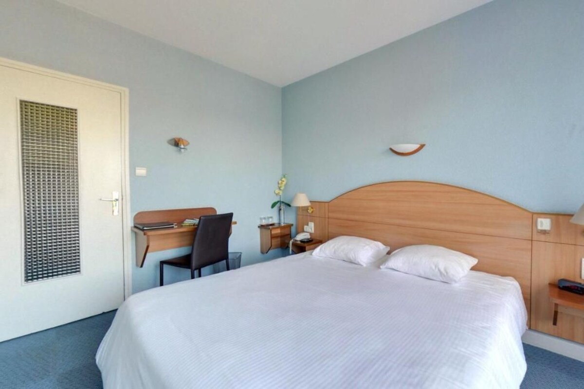 A hotel room with a bed, desk, chair, nightstand, lamp, and a small shelf.