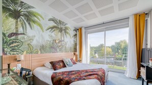 A hotel room with a bed, a desk, a chair, a window with curtains, and a mural of tropical plants on the wall. - Logis Hôtel la Maison Carrée (Mereville)