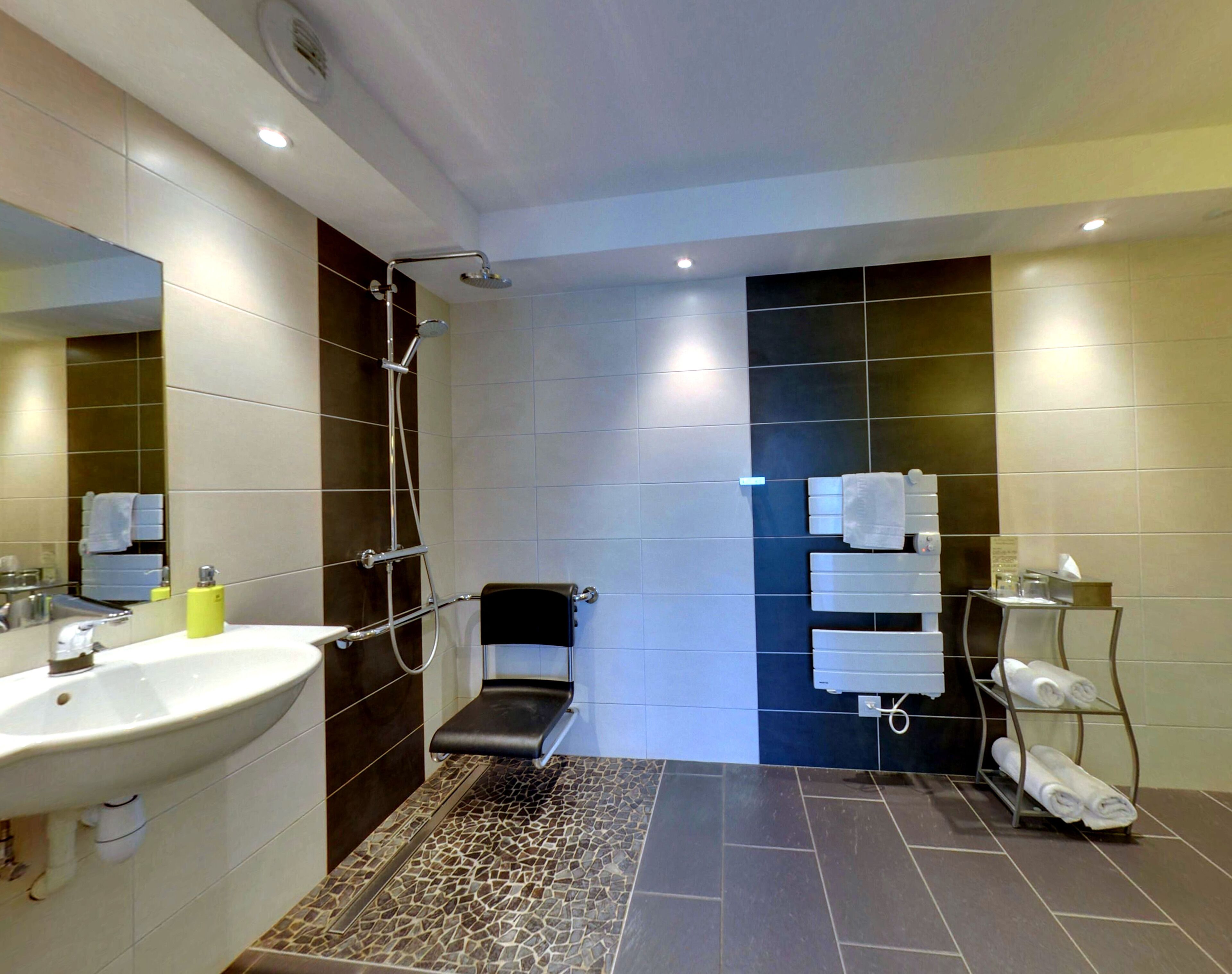 A modern bathroom with a large sink, a shower with a handheld showerhead, a bench, and a towel rack with towels.