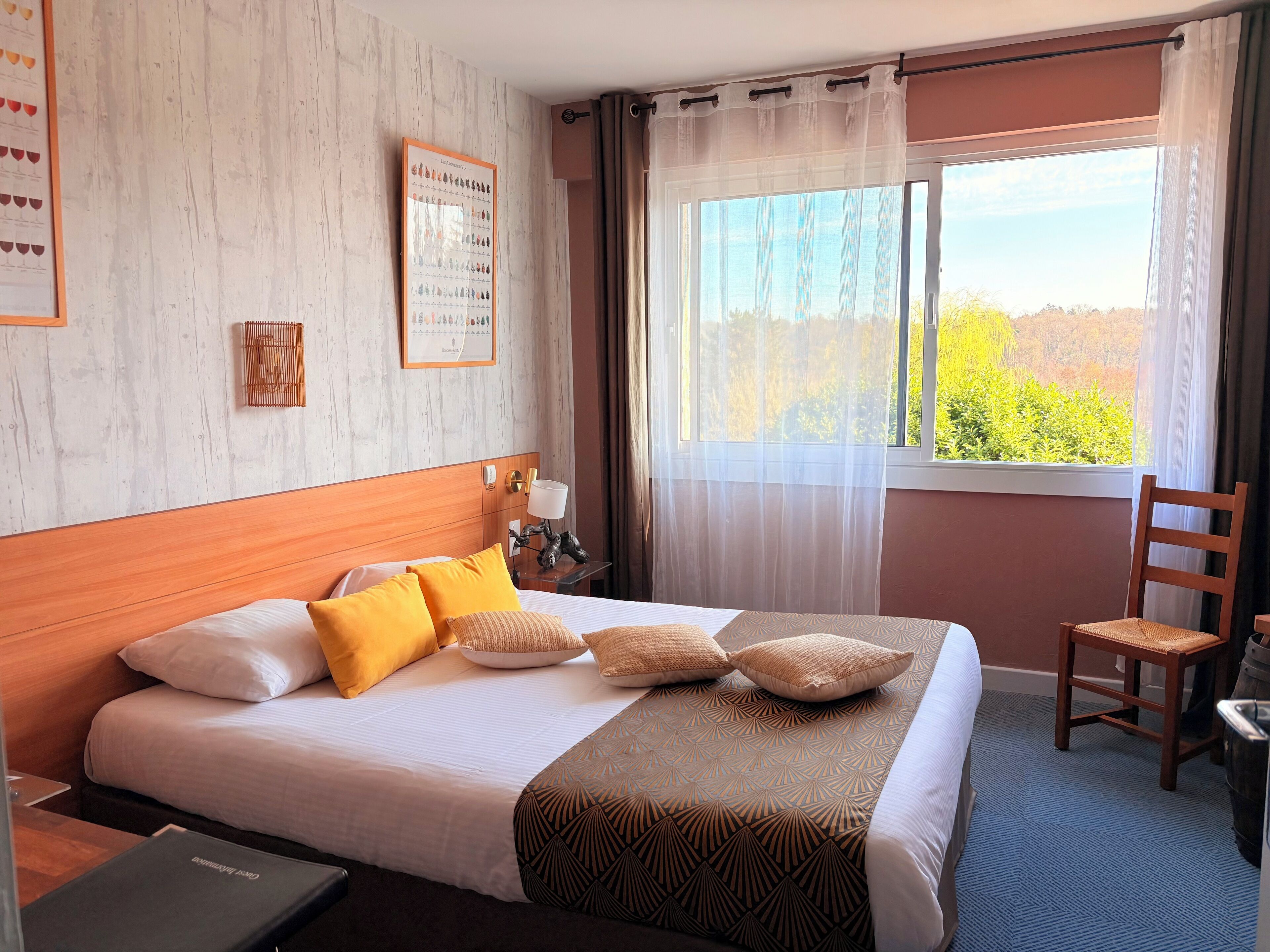 Hotelkamer met een groot bed, een houten hoofdeinde, een nachtkastje met een lamp, een stoel en een raam met uitzicht op bomen.