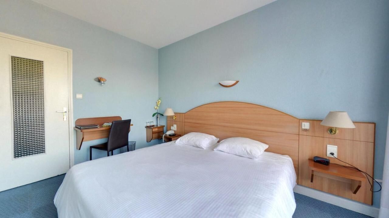 Et hotelværelse med seng, skrivebord, stol, natbord, lampe og en lille hylde.