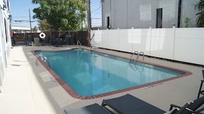 Piscina retangular com um deck branco e uma borda de tijolos. Há cadeiras de descanso e uma mesa pequena perto da piscina.