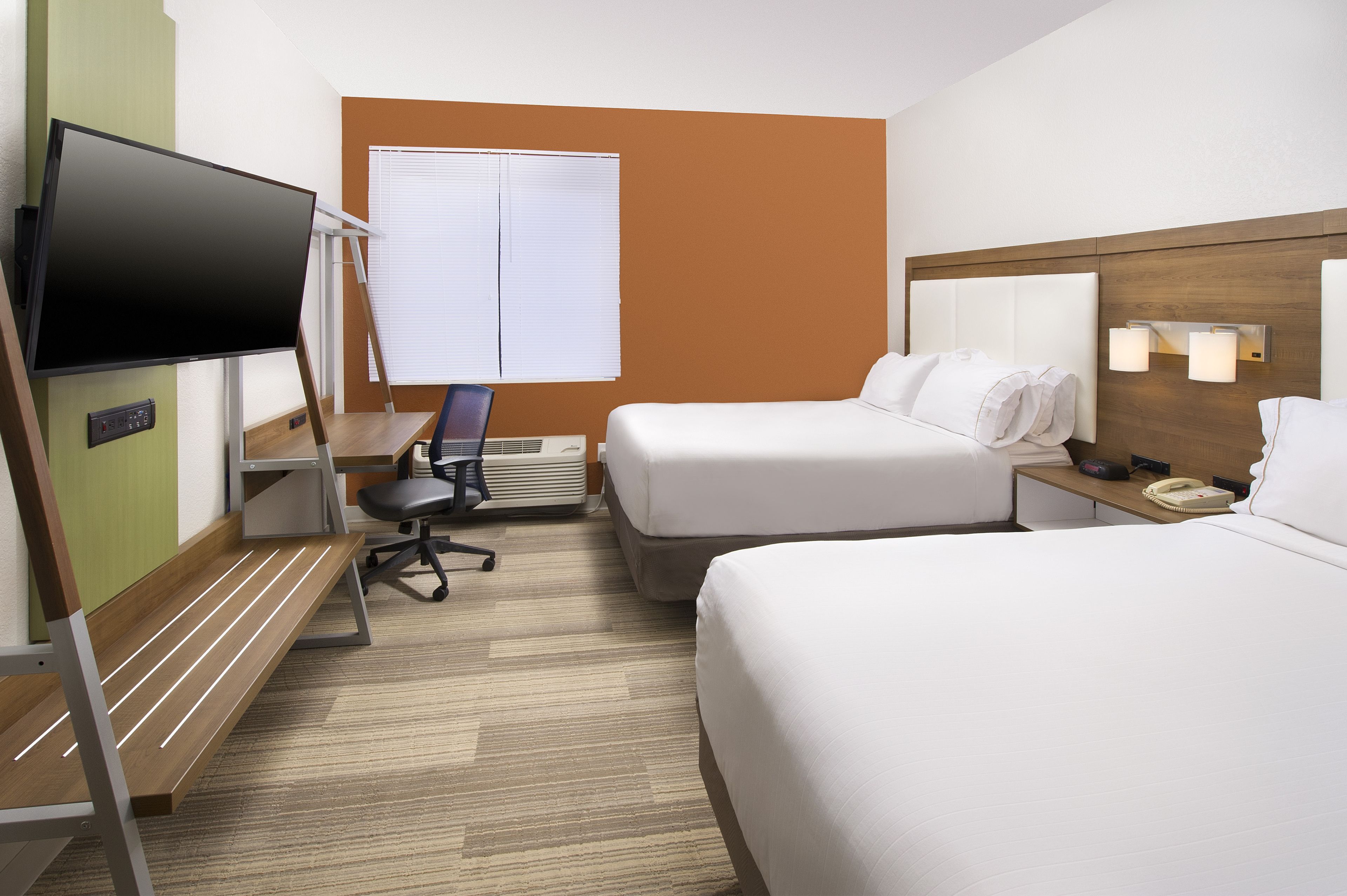 A hotel room with two beds, a TV, a desk with a chair, a window with blinds, and a bedside table with a lamp.