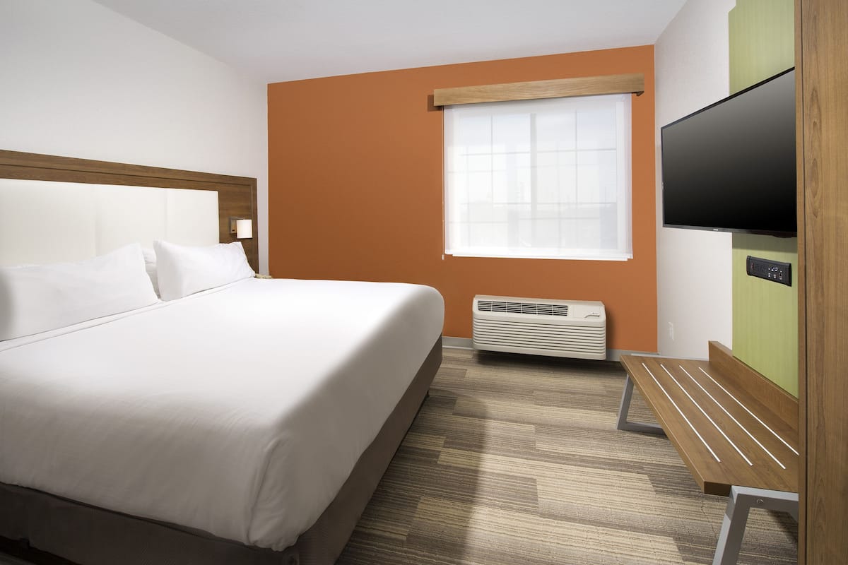 A hotel room with a large bed, a flat-screen TV, a window with blinds, a heater, and a wooden bench.