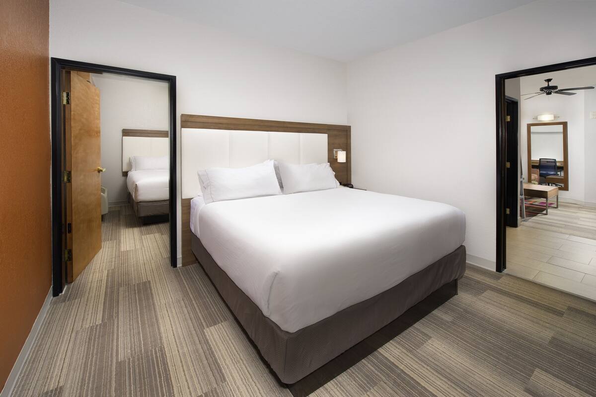 A hotel room with a large bed, a wooden headboard, a nightstand, a mirror, and a door leading to another room.