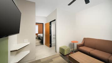 Une chambre d’hôtel équipée d’un téléviseur à écran plat, d’un canapé marron, d’une petite table ronde, d’un pouf à motifs et d’un couloir menant à la salle de bain.