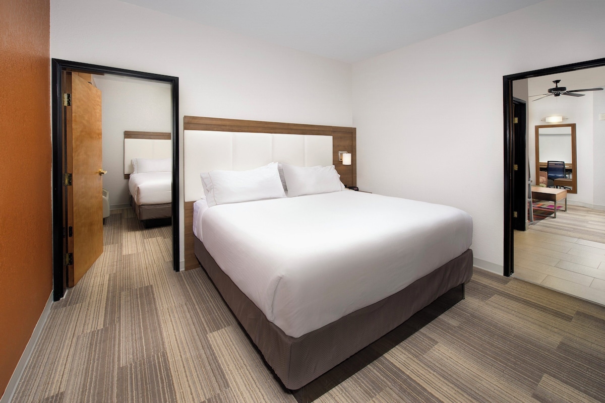 Quarto de hotel com uma cama grande, cabeceira de madeira, mesa de cabeceira, espelho e uma porta que dá para outro cômodo.