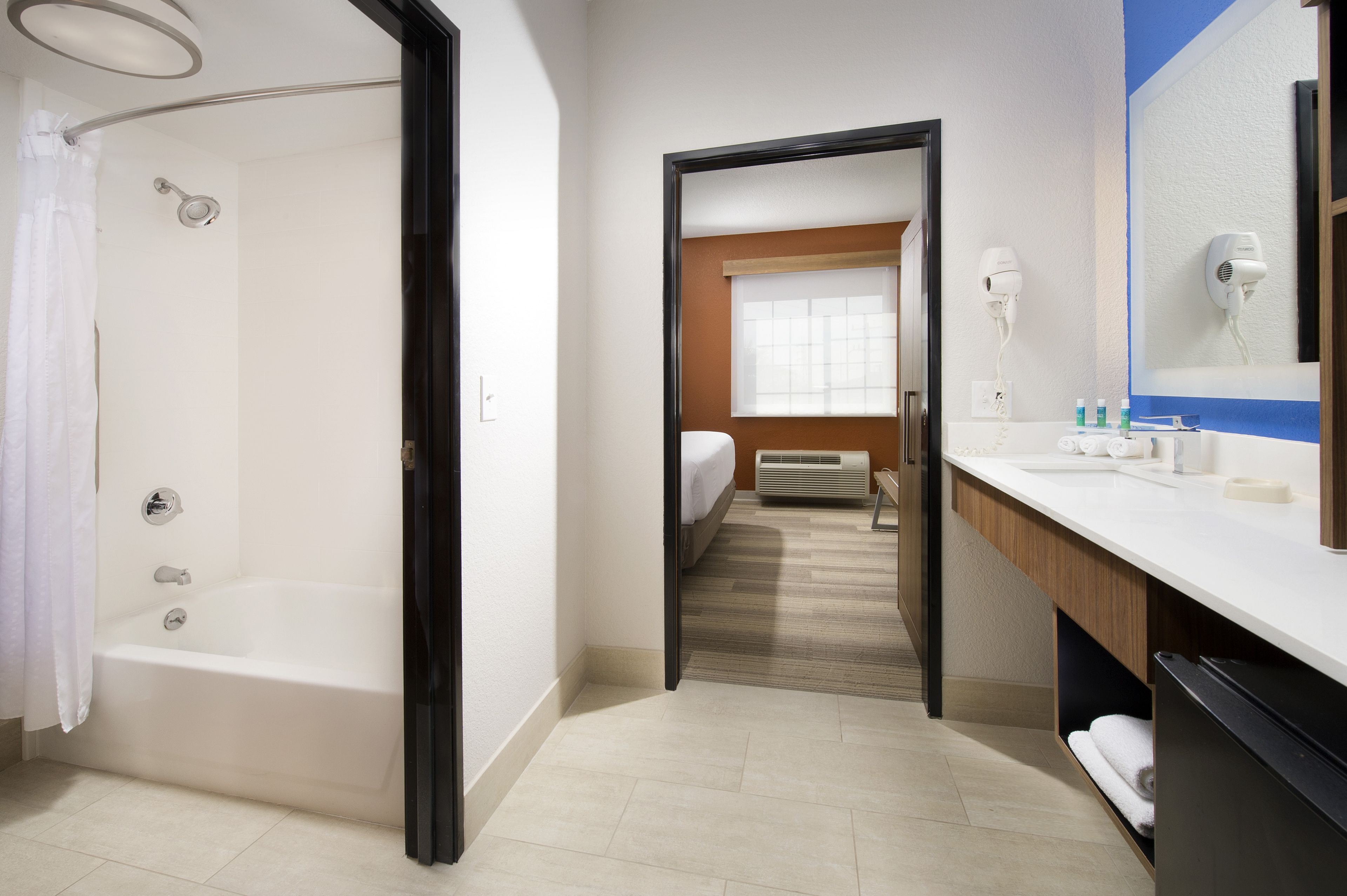 Une salle de bain moderne équipée d’une baignoire, d’une douche, d’un meuble-lavabo avec un miroir et d’un sèche-cheveux fixé au mur.