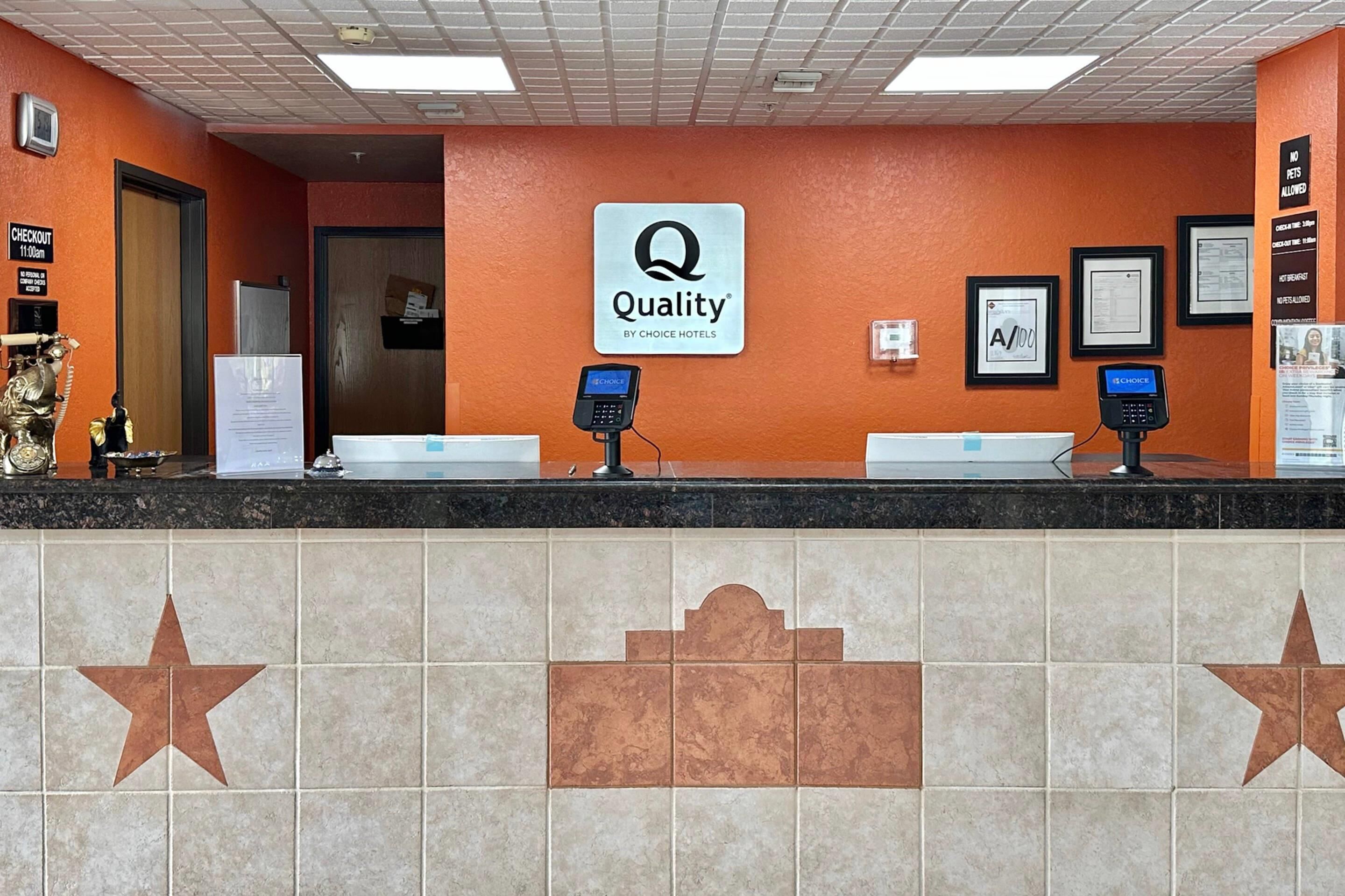 Un hall d'accueil d'hôtel avec une enseigne « Quality by Choice Hotels », deux comptoirs d'accueil, une enseigne « Départ » et des certificats encadrés accrochés au mur.