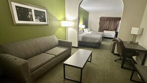 Een hotelkamer met een bank, een klein tafeltje, een bureau en een bed. Er staat een lamp op het bureau en nog een aan de muur. De kamer heeft groene wanden en een vloerkleed.