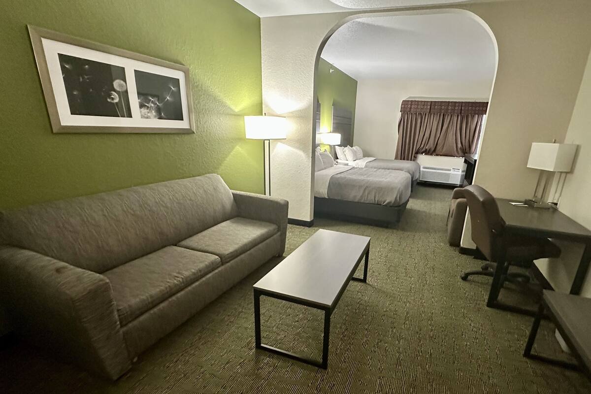 Een hotelkamer met een bank, een klein tafeltje, een bureau en een bed. Er staat een lamp op het bureau en nog een aan de muur. De kamer heeft groene wanden en een vloerkleed.