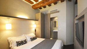 A modern hotel room with a large bed, bedside lamps, a wooden ceiling beam, a TV, and a bathroom with a glass shower enclosure. - Villa Des Princes (Paris)