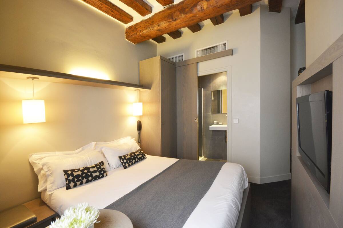 A modern hotel room with a large bed, bedside lamps, a wooden ceiling beam, a TV, and a bathroom with a glass shower enclosure.