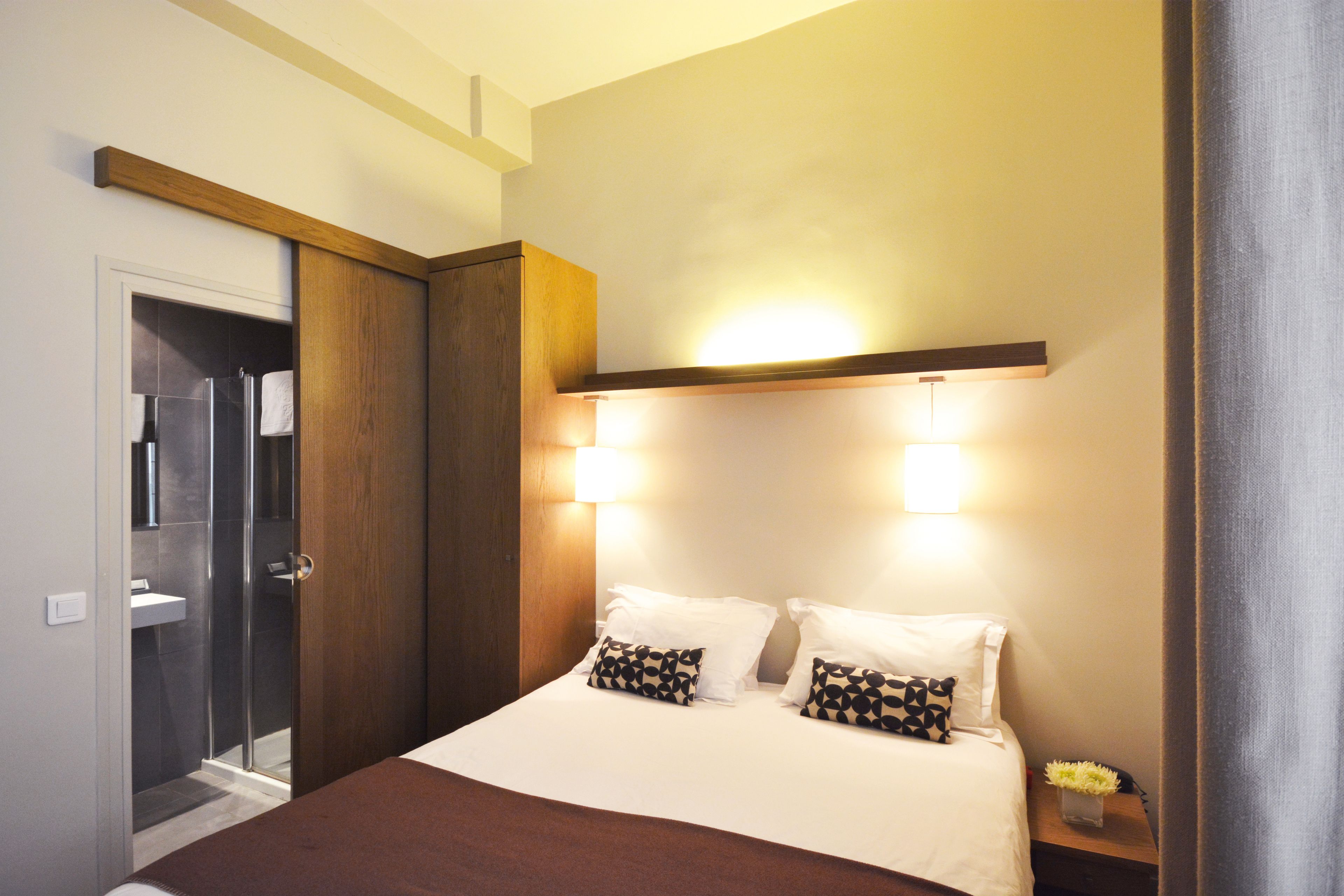 A hotel room with a bed, bedside lamps, a nightstand, a wooden wardrobe, and a shower area.