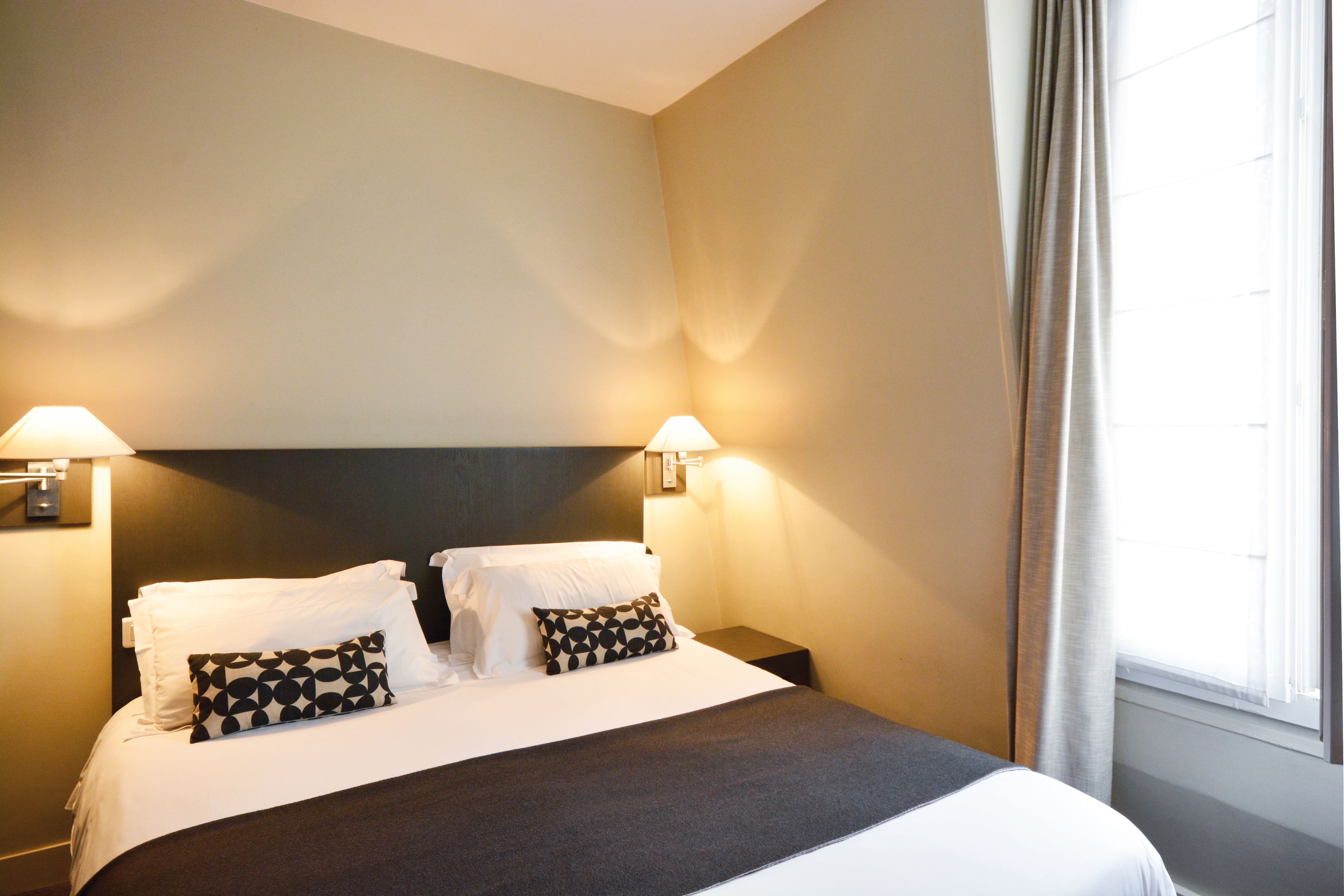 A neatly made bed with white linens and patterned pillows, two bedside lamps, and a window with curtains.