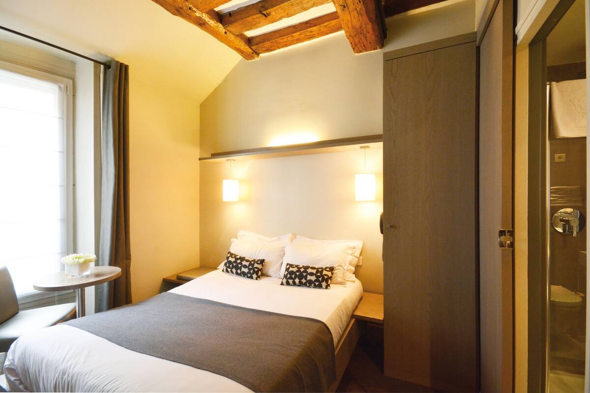 A hotel room with a bed, bedside tables, a small table with a vase, and a window with curtains.