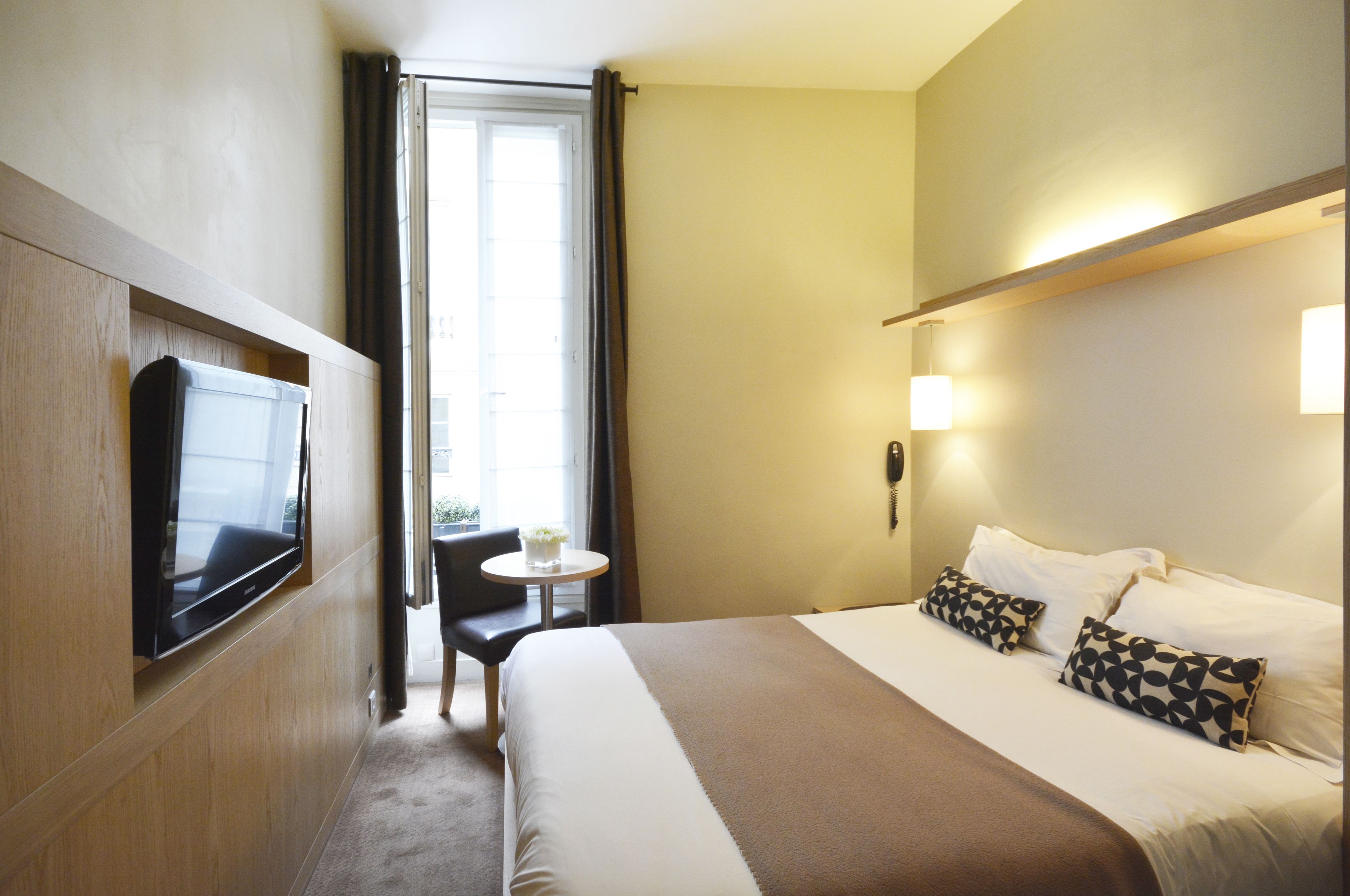 A hotel room with a bed, a TV mounted on the wall, a small table with a chair, and a window with curtains.
