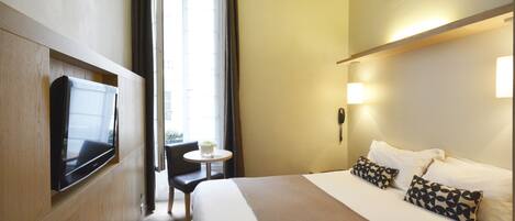 A hotel room with a bed, a TV mounted on the wall, a small table with a chair, and a window with curtains.
