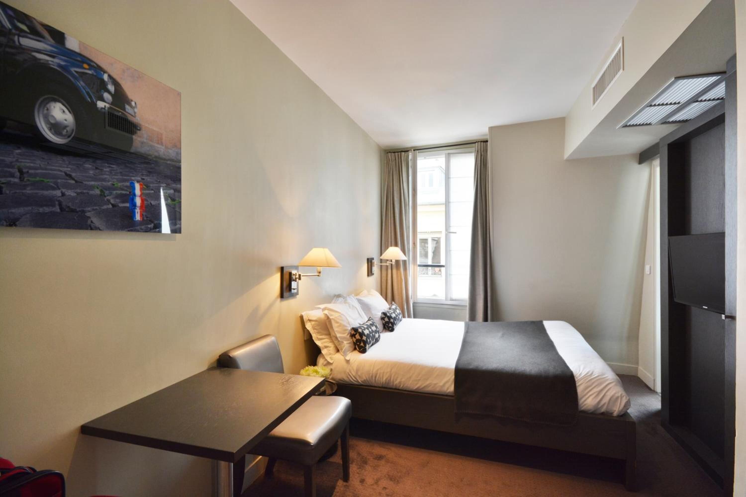 A hotel room with a bed, a desk, a chair, a lamp, a painting on the wall, and a window with curtains.