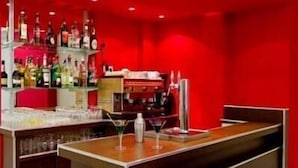 A bar area with a red wall, a wooden counter, and shelves stocked with bottles. - Hotel St-Etienne (Lourdes)
