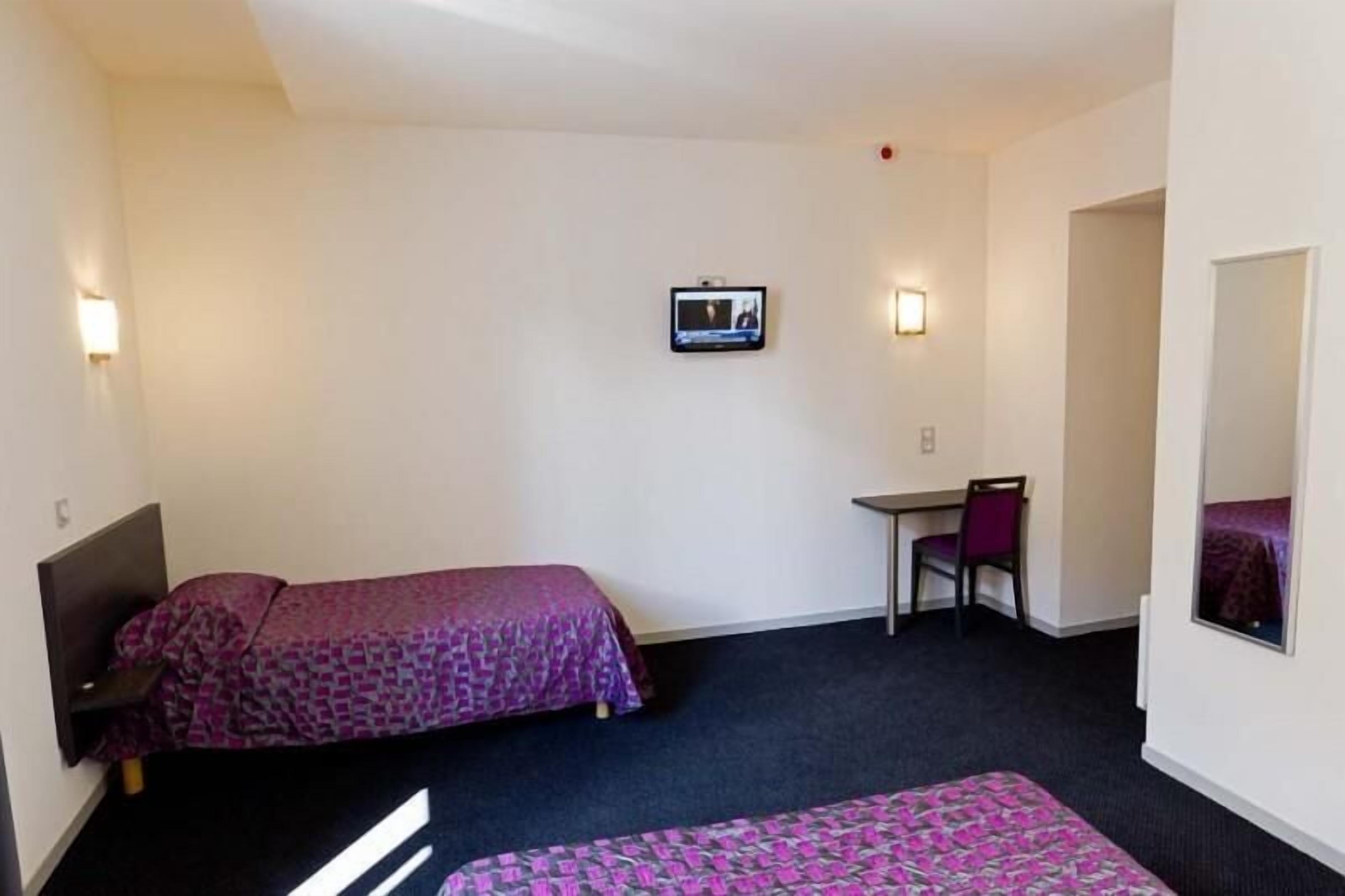 Ein kleines Hotelzimmer mit zwei Betten, einem Schreibtisch, einem Stuhl, einem an der Wand befestigten Fernseher und einem Spiegel.