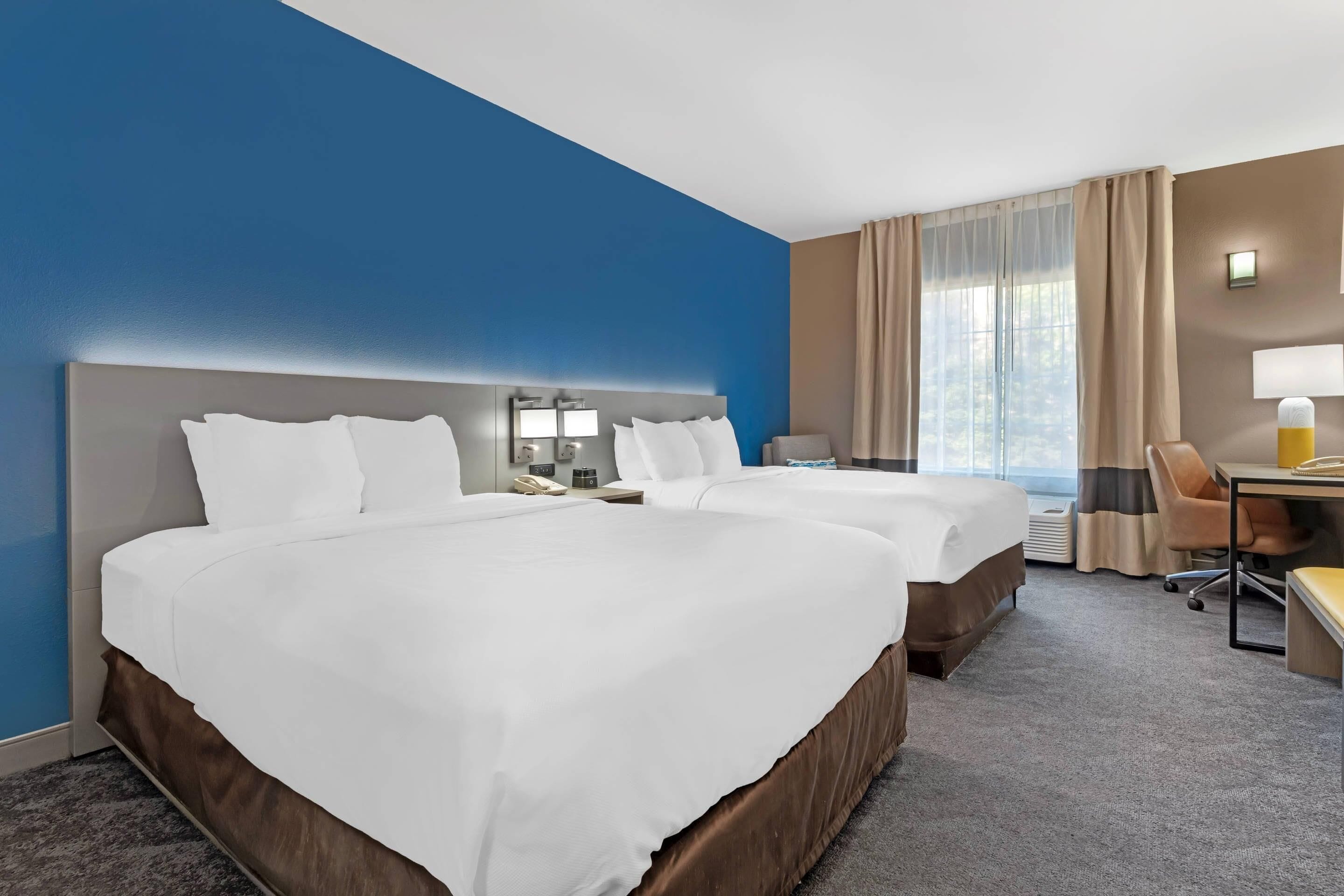Habitación de hotel con dos camas, un escritorio, una silla, una lámpara y una ventana con cortinas.