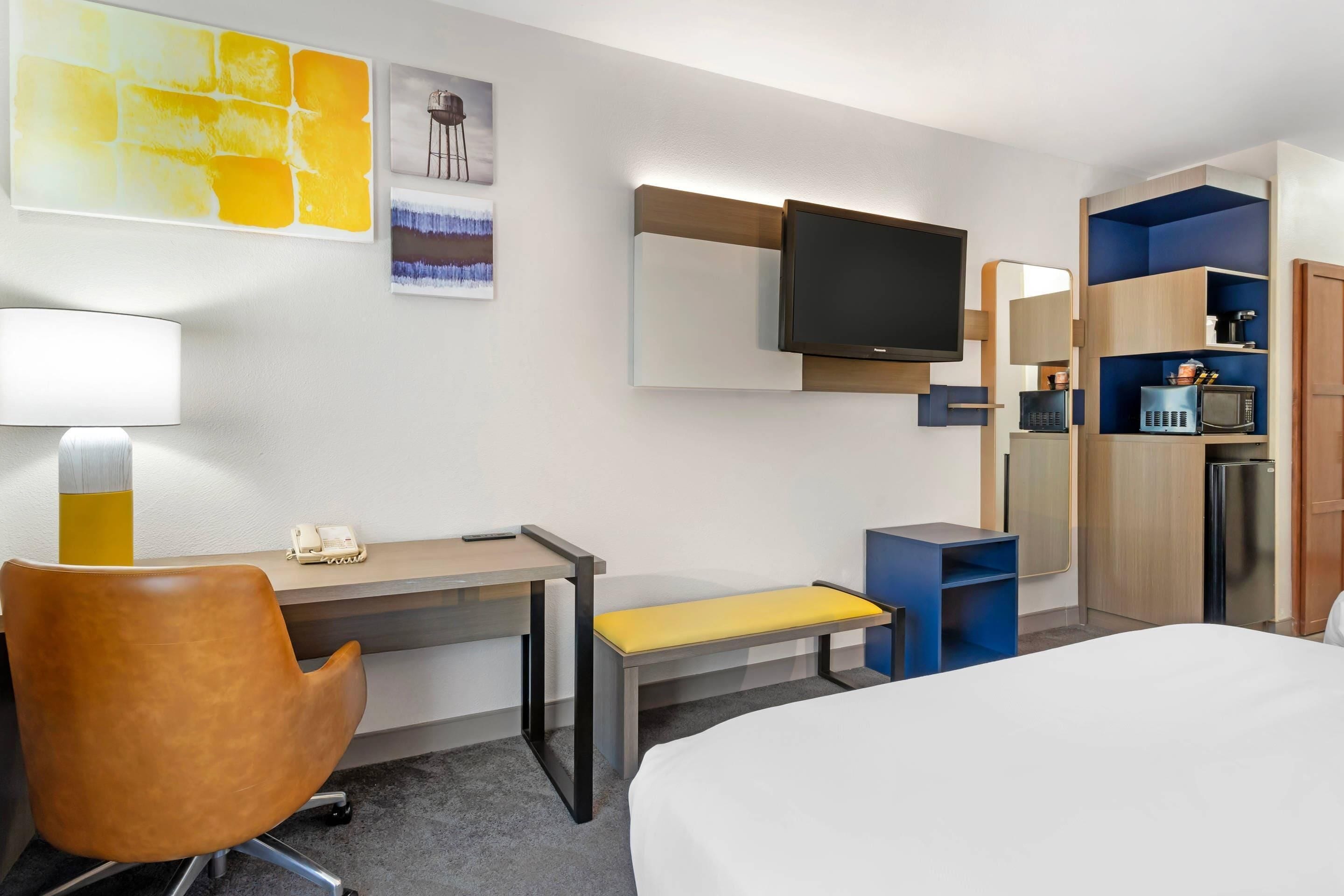A hotel room with a bed, desk, chair, bench, TV, and built-in kitchen area.