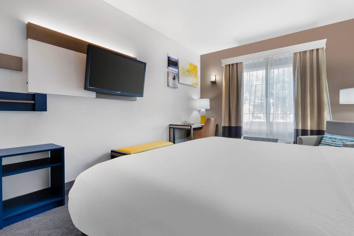A hotel room with a large bed, a TV mounted on the wall, a desk with a chair, and a window with curtains.