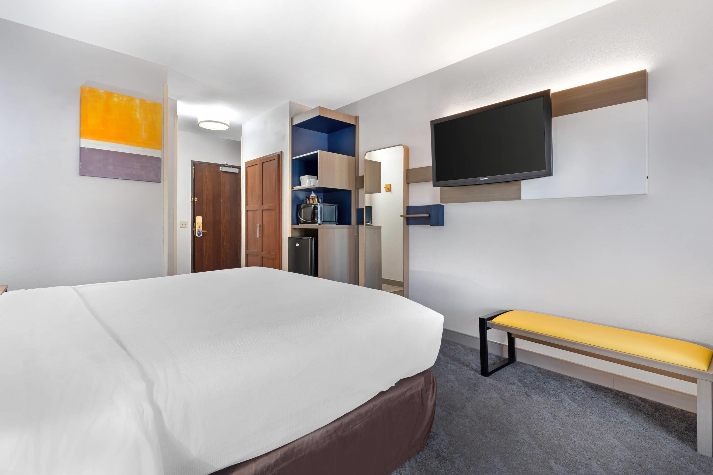 A hotel room with a large bed, a bench, a TV mounted on the wall, and a kitchenette area.
