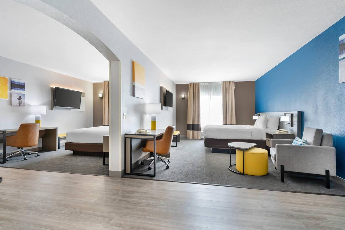 A modern hotel room with a bed, desk, chair, and a small seating area with a yellow ottoman.