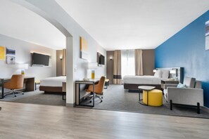 Una habitación de hotel moderna con cama, escritorio, silla y un pequeño rincón de descanso con un pouf amarillo.
