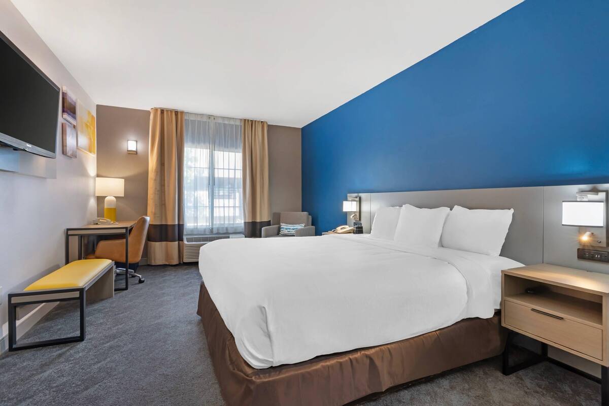 A hotel room with a large bed, a desk with a chair, a small table, a bench, and a TV mounted on the wall.