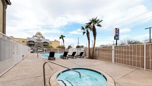 Área de hidromassagem com espreguiçadeiras e piscina, cercada por uma grade, com um hotel ao fundo.