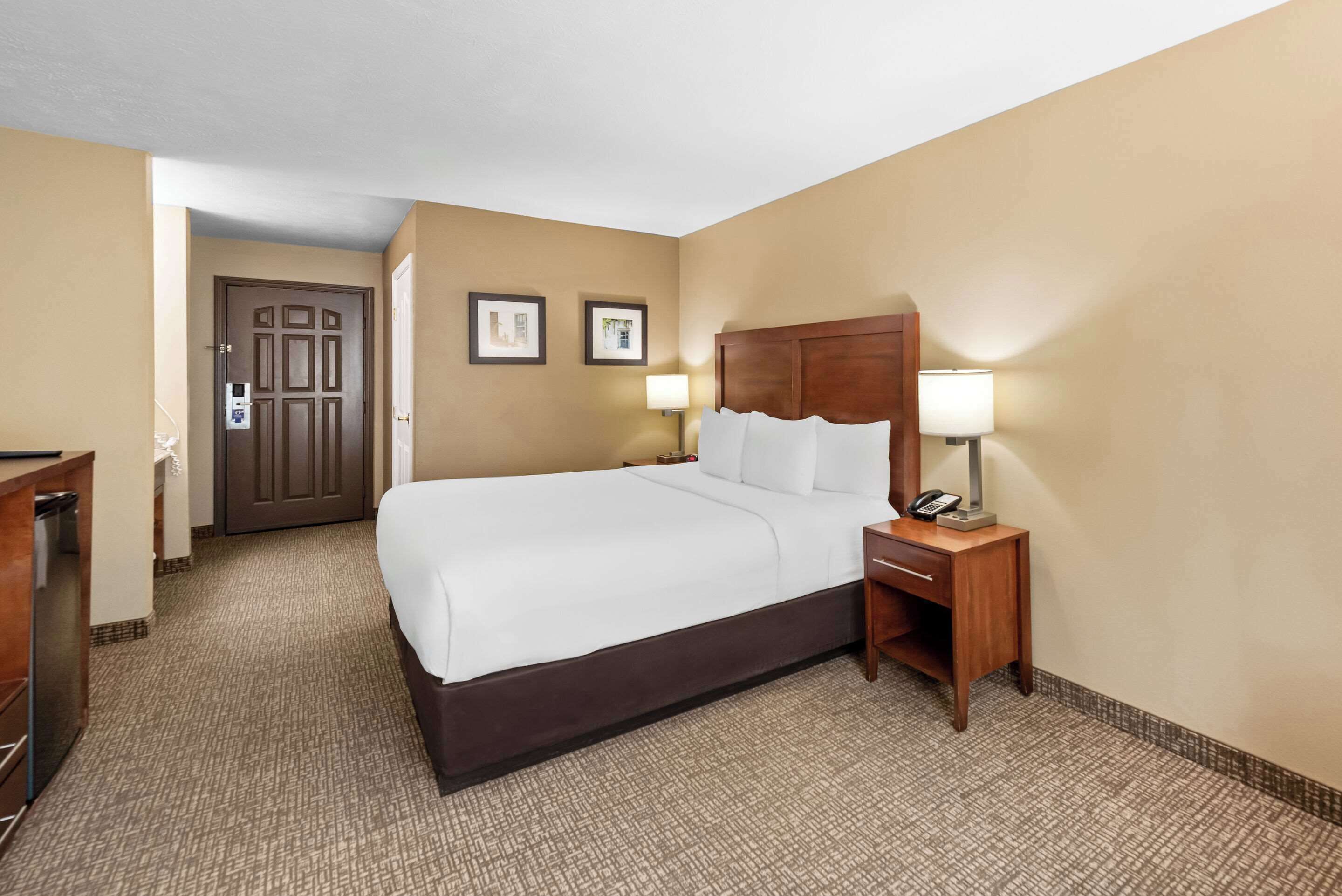 Una habitación de hotel con una cama grande, dos mesitas de noche con lámparas, un cabecero de madera y una puerta que da a otra habitación.