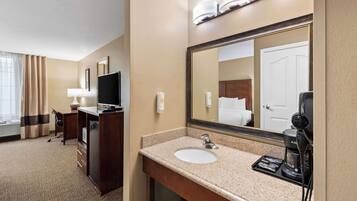 A hotel bathroom with a large mirror, a sink, a coffee maker, a desk, and a television.