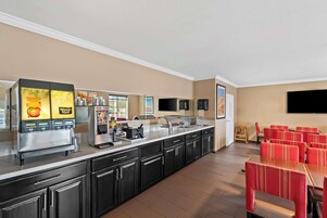 Un comedor de hotel con una estación de autoservicio para bebidas, un refrigerador y un televisor de pantalla plana montado en la pared.