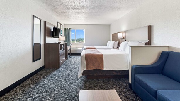 Habitación de hotel con una cama grande, una cama más pequeña, un escritorio con silla, un televisor montado en la pared, un sofá azul y una ventana con vistas al exterior.