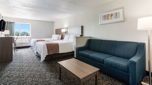Habitación de hotel con dos camas, un sofá azul, una mesa de centro de madera y un ventanal con vistas.