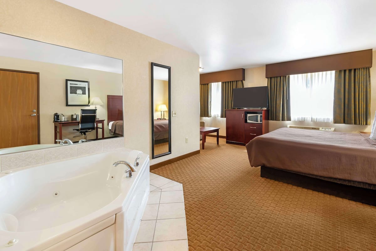 A hotel room with a large bed, a separate bathroom with a bathtub, a desk with a chair, a television, and a large mirror.