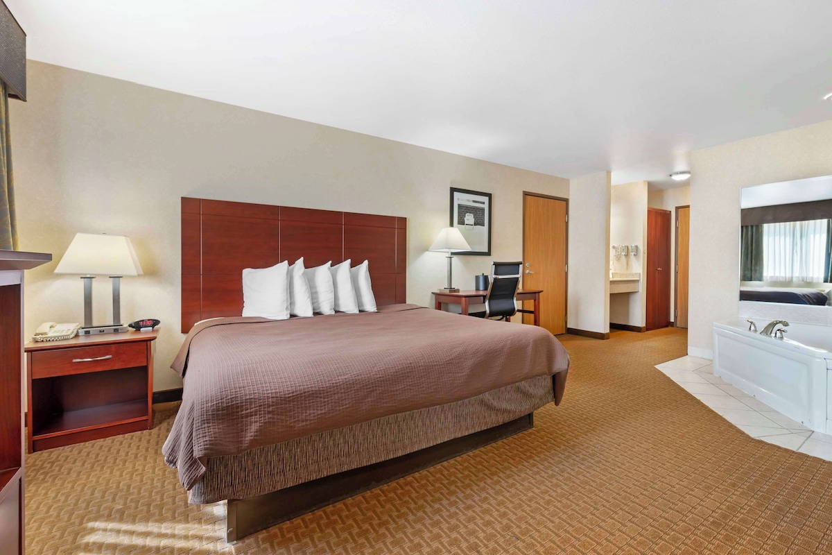 A hotel room with a large bed, a desk with a lamp, a chair, a bathroom with a bathtub, and a mirror.